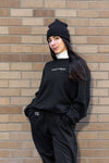 Goblin Suit: Ultra Cozy set shown against brick wall, featuring black fleece crew with white embroidered text and matching pants, styled with black beanie and white turtleneck. Cozy loungewear displayed in casual, comfortable setting.