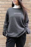 Goblin Suit: Ultra Cozy sweatshirt shown in dark grey with white Chaos Goblin embroidery across chest, styled with white turtleneck underneath against brick wall background, demonstrating comfortable oversized fit and premium fleece material.