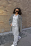 Ultra Cozy Open Leg Pant styled with a matching light grey sweatshirt and oversized tweed coat, showcasing the relaxed fit and casual elegance of the pants. White sneakers complete the monochromatic street style look against an urban brick backdrop.