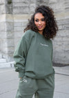 Stylish olive green Goblin Jogger set featuring a comfortable crewneck sweatshirt with embroidered logo and matching tapered jogger pants. Close-up view showing the cozy athleisure wear against a stone wall backdrop, highlighting the casual yet polished design and quality fabric texture.