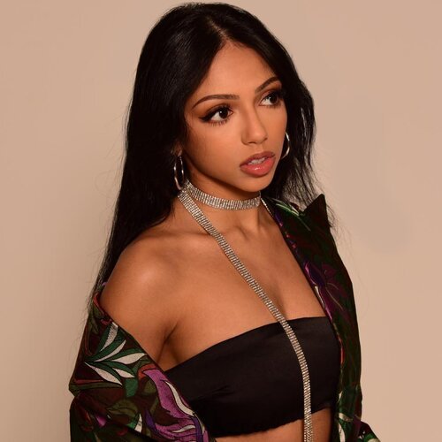 A press photo, headshot style of Saleka looking off to the right wearing a long necklace and black top and shirt with floral pattern off shoulder