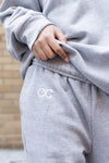 Ultra Cozy Open Leg Pant shown in light grey with CG embroidered logo on upper leg, demonstrating the relaxed fit and fleece-lined material. Close-up detail of the embroidered branding against the soft heather grey fabric texture.