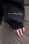 Team Tammy Crew Ultra Cozy black sweatshirt sleeve detail showing white embroidered text against brick wall background, showcasing sleeve embroidery and cozy fleece material.