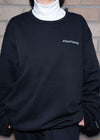 Team Tammy Crew: Ultra Cozy sweatshirt in black with white embroidered text on chest, shown with white turtleneck underneath. Close-up view displaying cozy fleece-lined material and classic crew neck design against brick wall background.