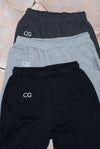 Ultra Cozy Open Leg Pant shown in three color variations - two black and one light grey sweatpants, each featuring CG embroidered logo on upper right leg, displayed in a layered arrangement against a light textured background.