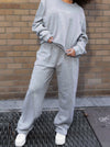 Ultra Cozy Open Leg Pant shown in light grey, featuring an oversized fit with fleece lining and CG embroidery on upper right leg. Styled with matching sweatshirt and white sneakers against brick wall background, demonstrating comfortable casual wear.