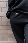 Ultra Cozy Open Leg Pant in dark grey with white CG embroidery detail shown against brick wall background, highlighting the relaxed fit and premium fleece material. Close-up view emphasizes the branded embroidery on upper right leg.