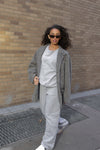 Ultra Cozy Open Leg Pant styled with a matching light grey sweatshirt and oversized tweed coat, showcasing the relaxed fit and casual elegance of the pants. White sneakers complete the monochromatic street style look against an urban brick backdrop.