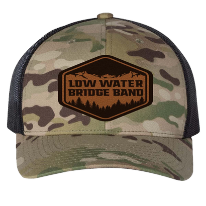 Multicam trucker hat with black mesh back, featuring a leather patch with Low Water Bridge Band logo and mountain scenery, structured six-panel design, and snapback closure