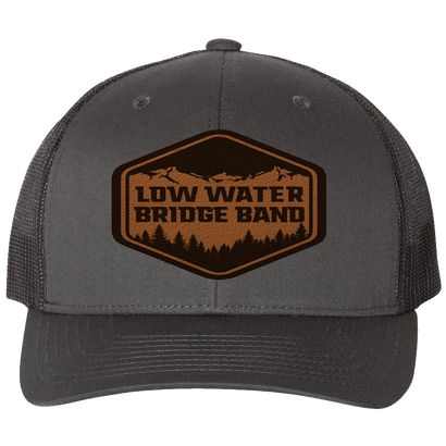 Charcoal trucker hat with black mesh back featuring brown leather patch with Low Water Bridge Band logo, mountain silhouette, and trees. Structured six-panel design with snapback closure.