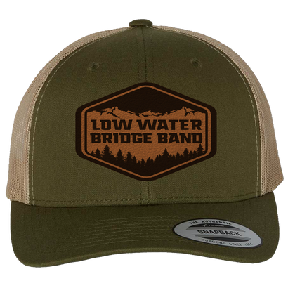 LWBB Leather Patch Trucker hat in moss green and khaki with faux leather patch featuring mountain scenery and forest silhouette. Structured mid-profile design with mesh back and snapback closure.