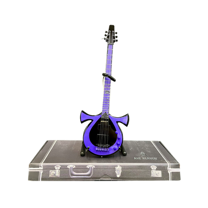 Miniature Oteil Burbridge Signature Ankh 6-string bass guitar replica in purple, displayed on stand atop a vintage-style case. Handcrafted 1:4 scale model with unique ankh-shaped body and detailed fretboard.