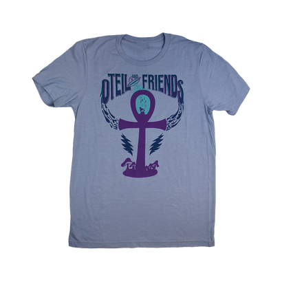 Light blue t-shirt featuring Oteil & Friends logo with purple ankh symbol, lightning bolt wings, and band name in stylized text. Design combines Egyptian symbolism with rock music imagery on a comfortable unisex tee.