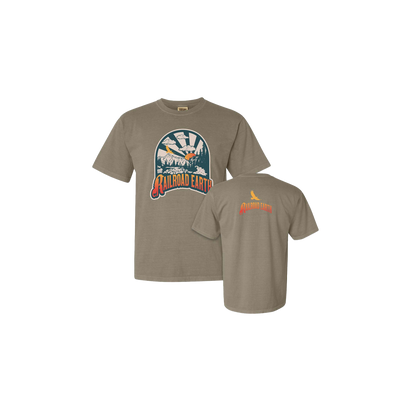 River Tee in khaki featuring a circular mountain river graphic with sunburst design and bird silhouette. Front displays scenic outdoor illustration while back shows small brand logo in orange. Comfort Colors unisex t-shirt in earthy tan shade.